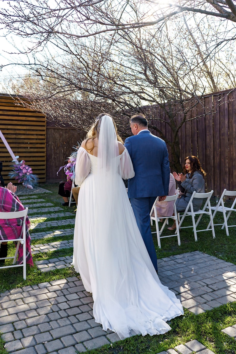 May include: A bride in a white wedding dress and veil walks down the aisle with her father. Guests are seated in white chairs, watching the ceremony.