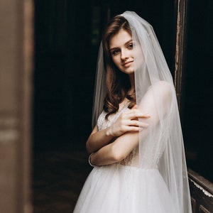 May include: A bride wearing a white wedding dress with a long veil, standing in a doorway. She is looking at the camera and smiling.