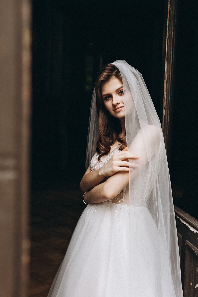 May include: A bride wearing a white wedding dress with a long veil, standing in a doorway. She is looking at the camera and smiling.