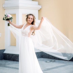 May include: A bride in a white wedding dress with a long veil, holding a bouquet of flowers. She is standing in front of a building with a white column.
