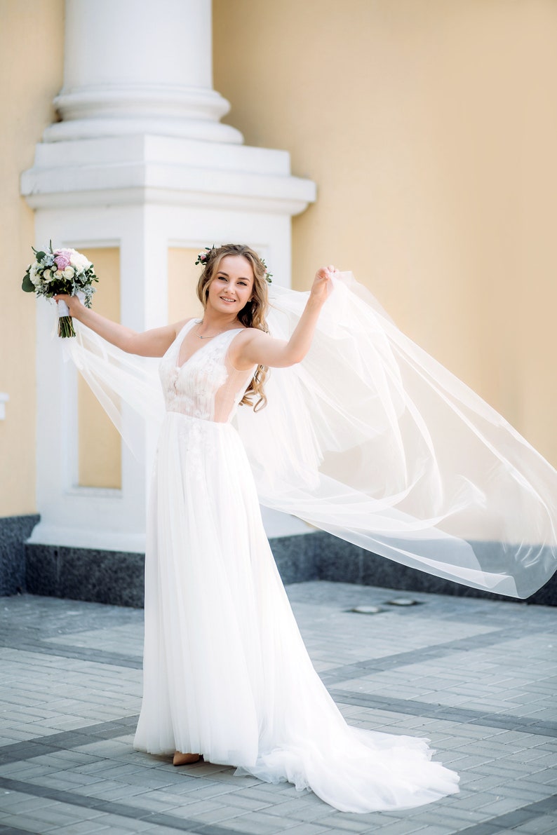 May include: A bride in a white wedding dress with a long veil, holding a bouquet of flowers. She is standing in front of a building with a white column.
