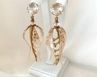 May include: A pair of elegant, gold-toned earrings. Each earring features a large, clear, oval-shaped stone at the top, connected to a detailed, openwork design with a floral or botanical theme. The earrings are displayed on white stands.