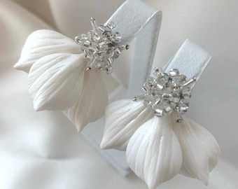May include: A pair of white floral earrings. Each earring features three textured petals and a cluster of small, clear beads at the center. The earrings are displayed on a white stand against a white background.