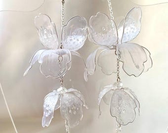 May include: Elegant, clear, floral-shaped earrings. Each earring features two large, translucent flower designs with delicate silver wire detailing and small, dangling crystal accents. The earrings have a sparkling, ethereal quality.
