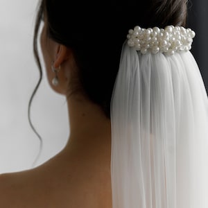 Puede incluir: Accesorio para el cabello de novia con un grupo de perlas de imitaci&oacute;n. Las perlas est&aacute;n unidas a un peine, con un velo blanco transparente en cascada. La imagen muestra la espalda de una persona, con el accesorio para el cabello y el velo enfocados.