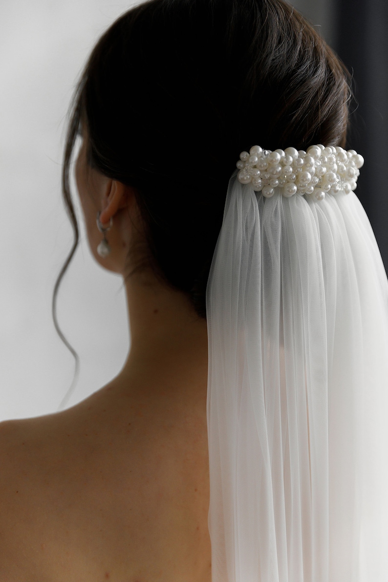 Puede incluir: Accesorio para el cabello de novia con un grupo de perlas de imitaci&oacute;n. Las perlas est&aacute;n unidas a un peine, con un velo blanco transparente en cascada. La imagen muestra la espalda de una persona, con el accesorio para el cabello y el velo enfocados.