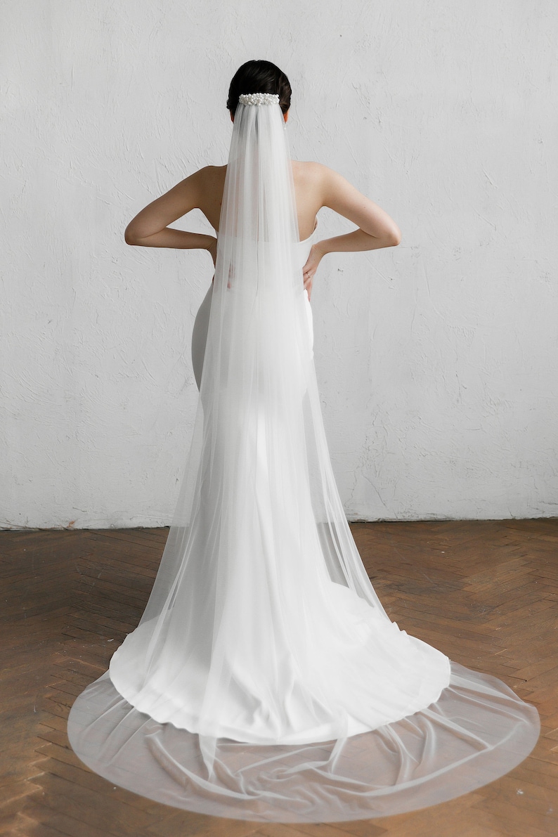 Puede incluir: Un velo de novia blanco con un adorno para la cabeza con cuentas. El velo es largo y fluido, extendi&eacute;ndose hasta el suelo. La novia lleva un vestido blanco. El fondo es una pared blanca y un suelo de madera.