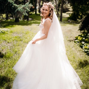 May include: A bride in a white wedding dress with a long veil, standing in a grassy field with trees in the background.