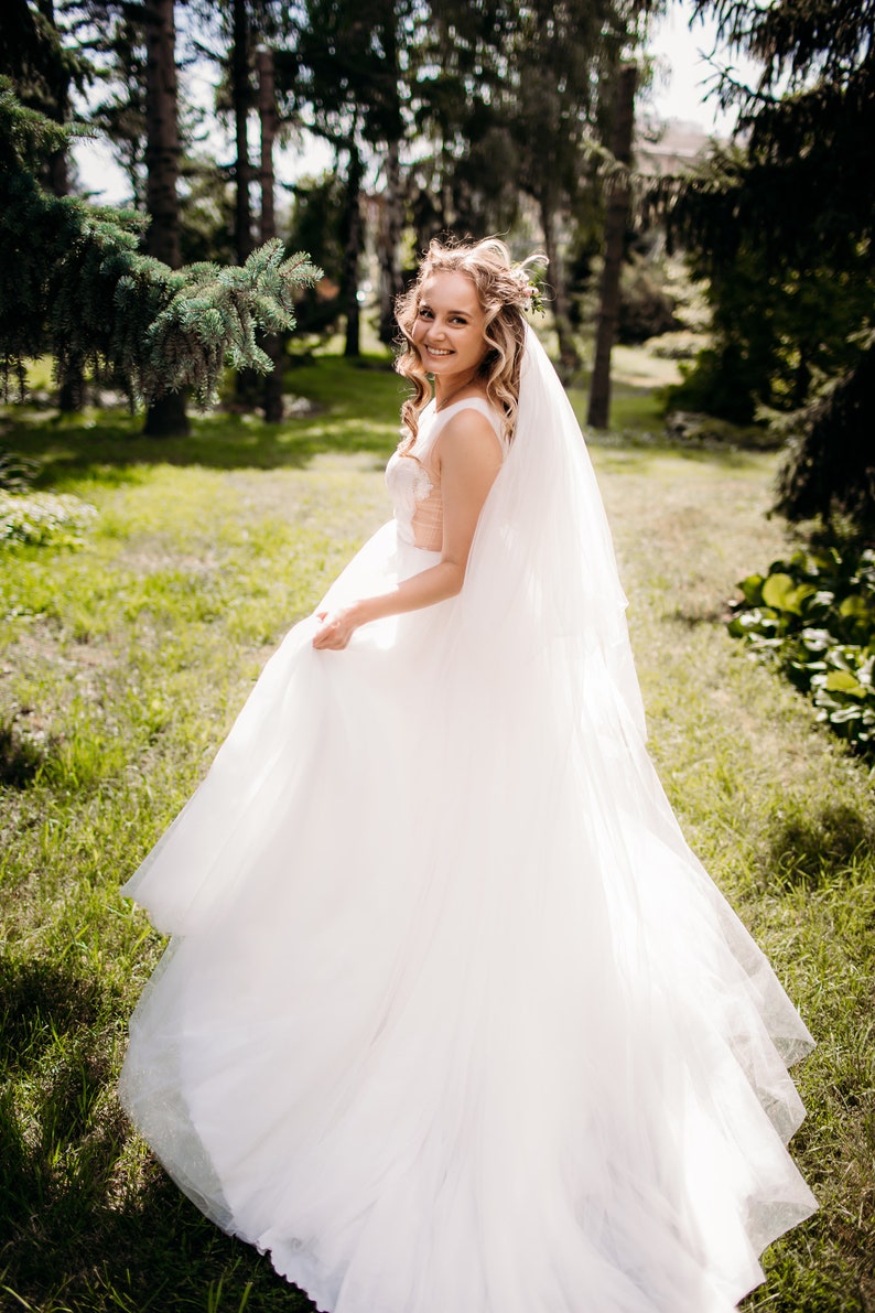 May include: A bride in a white wedding dress with a long veil, standing in a grassy field with trees in the background.