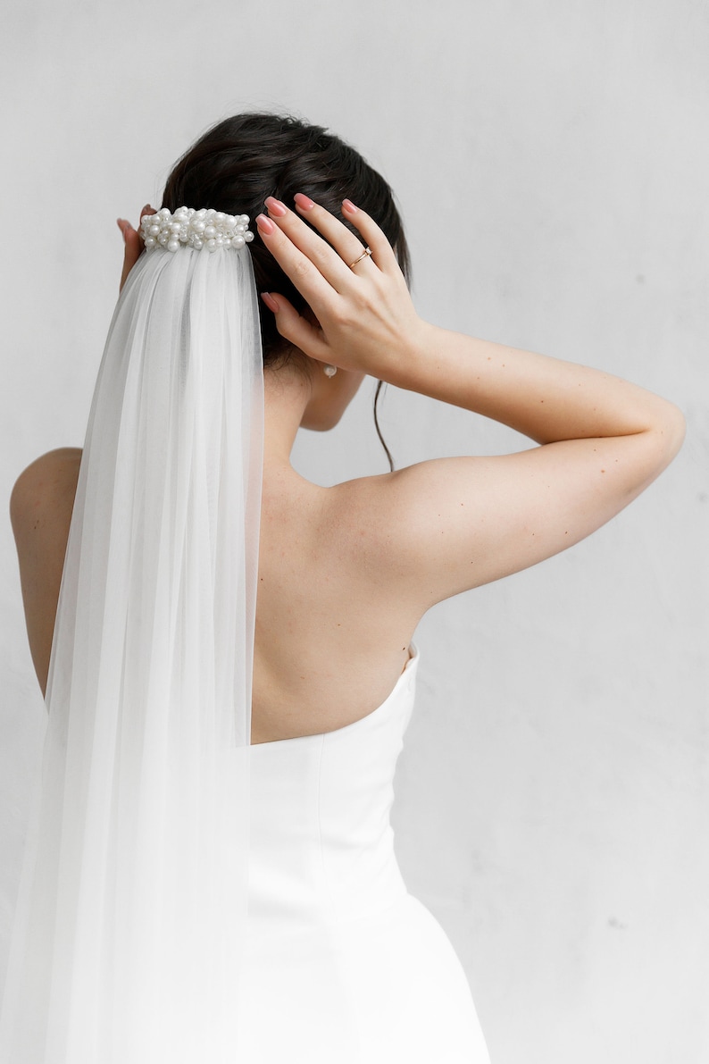 Puede incluir: Peinado de novia con un adorno para el cabello adornado con perlas y un velo blanco transparente. La novia lleva un vestido blanco. La imagen est&aacute; tomada desde atr&aacute;s, mostrando la parte posterior de la cabeza y los hombros de la novia.