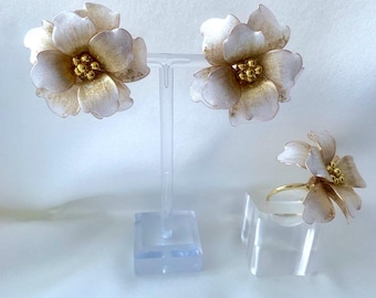 May include: A floral jewelry set featuring a pair of earrings and a ring. The earrings and ring are shaped like flowers with white petals and gold centers. The jewelry is displayed on clear stands against a white background.