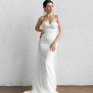 Puede incluir: Un vestido de novia blanco sin tirantes con escote coraz&oacute;n y cola fluida. El vestido es entallado y est&aacute; hecho de una tela lisa, similar al sat&eacute;n. Un velo blanco transparente cae por la espalda.