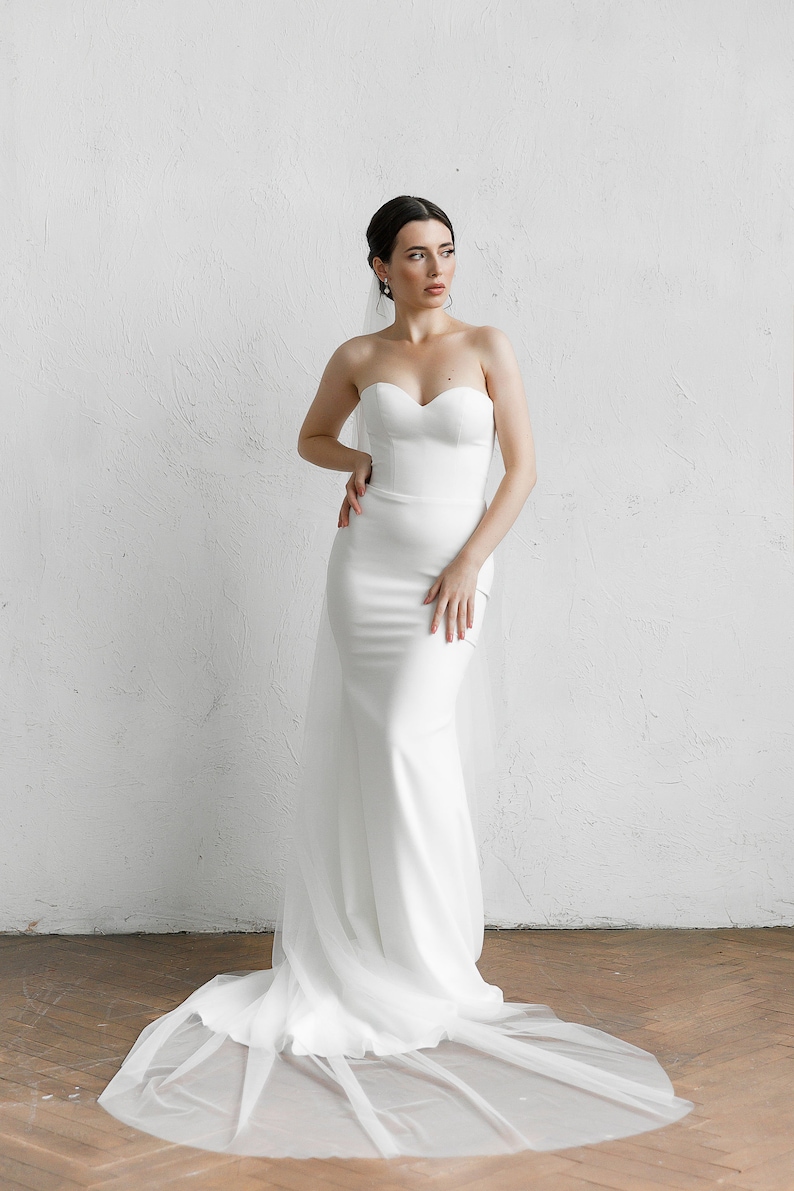 Puede incluir: Un vestido de novia blanco sin tirantes con escote coraz&oacute;n y cola fluida. El vestido es entallado y est&aacute; hecho de una tela lisa, similar al sat&eacute;n. Un velo blanco transparente cae por la espalda.