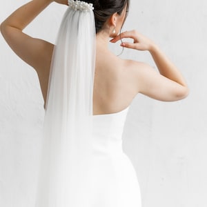 Puede incluir: Una novia con un vestido de novia blanco y velo. El velo est&aacute; sujeto a un adorno para el cabello adornado con perlas. La espalda de la novia est&aacute; hacia la c&aacute;mara, con los brazos levantados. El fondo es una pared blanca lisa.