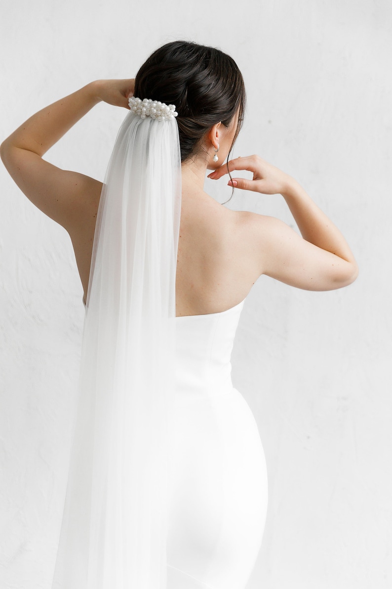 Puede incluir: Una novia con un vestido de novia blanco y velo. El velo est&aacute; sujeto a un adorno para el cabello adornado con perlas. La espalda de la novia est&aacute; hacia la c&aacute;mara, con los brazos levantados. El fondo es una pared blanca lisa.