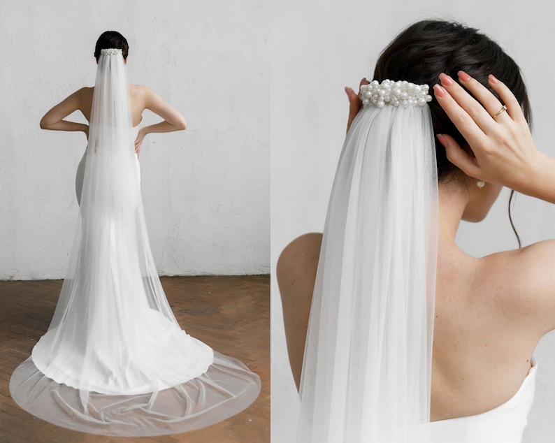 Puede incluir: Un velo de novia blanco con un dise&ntilde;o delicado y fluido. El velo est&aacute; sujeto a un peine para el cabello adornado con perlas. La novia lleva un vestido blanco. El velo cae por la espalda, creando un aspecto suave y elegante para una boda.