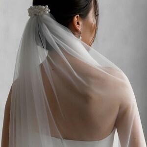 Puede incluir: Un velo de novia blanco y un accesorio para el cabello. El velo es transparente y cae por la espalda. Una horquilla para el pelo con perlas asegura el velo. La novia lleva un vestido blanco sin tirantes y pendientes de perlas.
