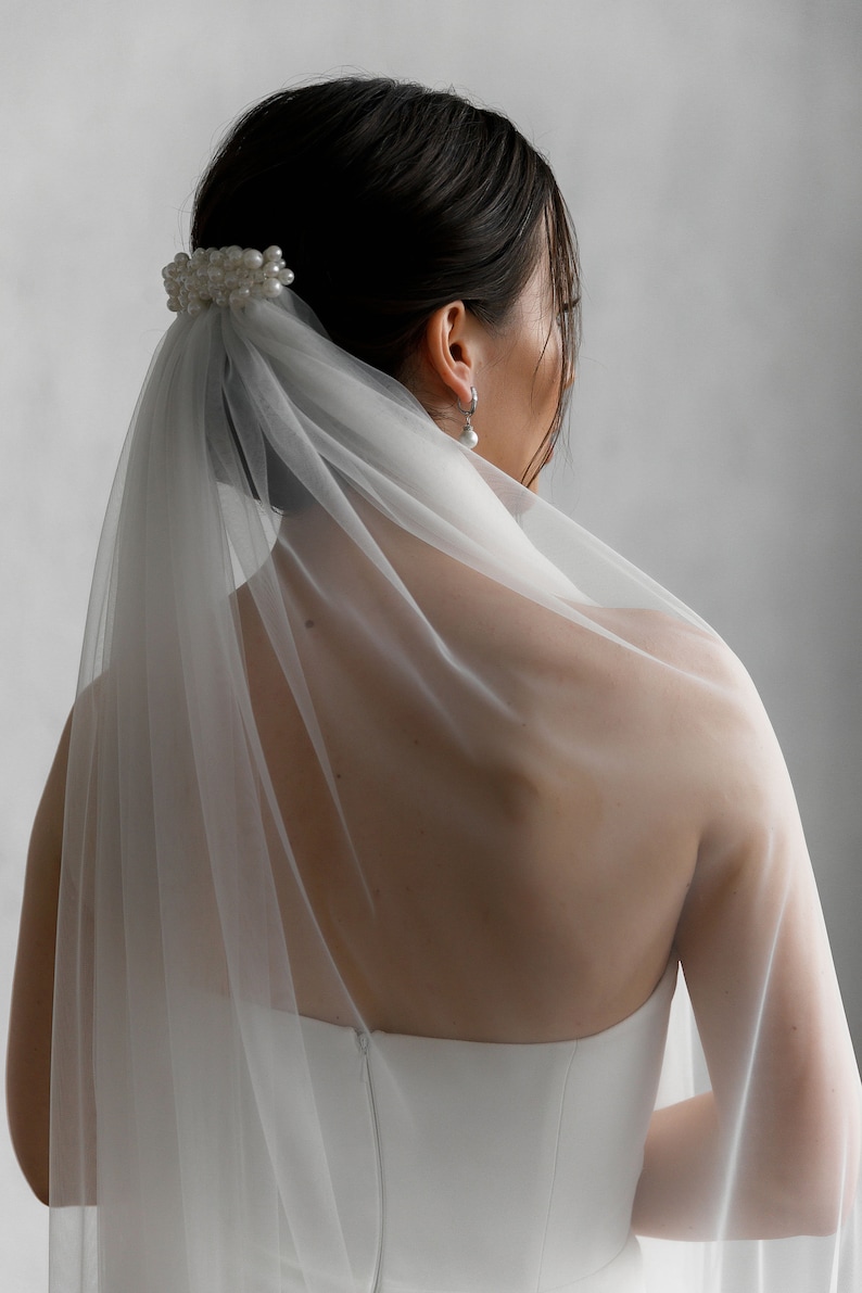 Puede incluir: Un velo de novia blanco y un accesorio para el cabello. El velo es transparente y cae por la espalda. Una horquilla para el pelo con perlas asegura el velo. La novia lleva un vestido blanco sin tirantes y pendientes de perlas.