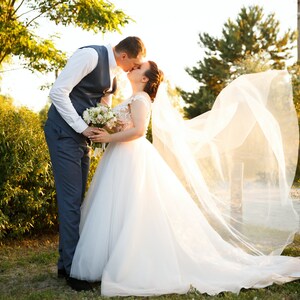 May include: A bride and groom kiss under a veil blowing in the wind. The bride is wearing a white wedding dress with a long veil. The groom is wearing a blue suit and a white shirt. They are standing in a grassy area with trees in the background.