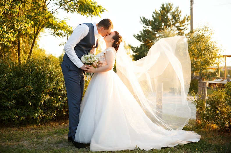 May include: A bride and groom kiss under a veil blowing in the wind. The bride is wearing a white wedding dress with a long veil. The groom is wearing a blue suit and a white shirt. They are standing in a grassy area with trees in the background.