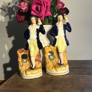 Könnte beinhalten: Zwei Porzellanfiguren von Männern in blauen Jacken und gelben Hosen, die auf gelben und braunen Sockeln stehen. Die Figuren halten Vögel in ihren Händen. Die Figuren stehen vor einer weißen Vase mit weißen und rosa Blumen.