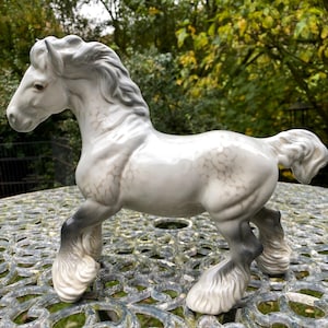 May include: A ceramic horse figurine, predominantly white with grey shading and black lower legs. The mane and tail are grey. The figurine has a glossy finish. It is posed as if walking. The background is a blurred outdoor scene with green foliage.
