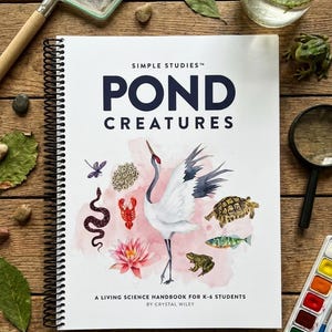 May include: A spiral-bound book titled "Pond Creatures" with illustrations of pond life, including a crane, turtle, and fish. The book is surrounded by a net, magnifying glass, and watercolor paints on a wooden surface.