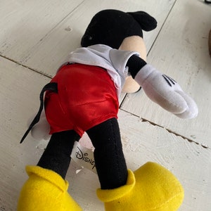 Może przedstawiać: Pluszowa zabawka Mickey Mouse w czerwonych satynowych spodenkach, białej koszuli, czarnych butach i czarnym ogonie. Zabawka ma przyczepioną do nogi metkę Disney.