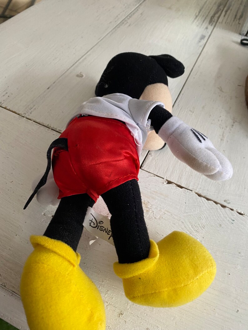 Może przedstawiać: Pluszowa zabawka Mickey Mouse w czerwonych satynowych spodenkach, białej koszuli, czarnych butach i czarnym ogonie. Zabawka ma przyczepioną do nogi metkę Disney.