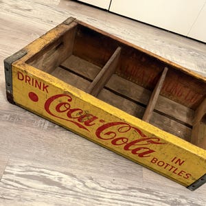 May include: Vintage Coca-Cola wooden crate with red lettering on a yellow background. The crate is divided into three sections and has metal corner reinforcements. The text reads "DRINK Coca-Cola IN BOTTLES".