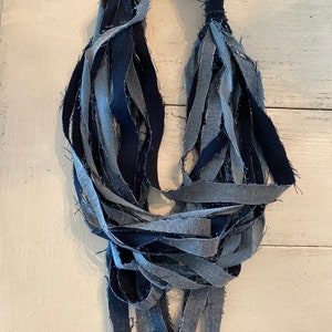 May include: A long necklace made from strips of blue denim fabric. The fabric is frayed and knotted together to create a unique and textured look.