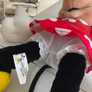 Może przedstawiać: Czerwono-biała sukienka w kropki z białym halką i czarnymi butami. Sukienka ma białą koronkową lam&oacute;wkę. Lalka nosi czarną opaskę na głowę z kokardką. Lalka ma ż&oacute;łtą twarz i czarne oczy. Lalka trzyma białą poduszkę w kształcie serca. Lalka ma metkę z napisem "Walt Disney World".