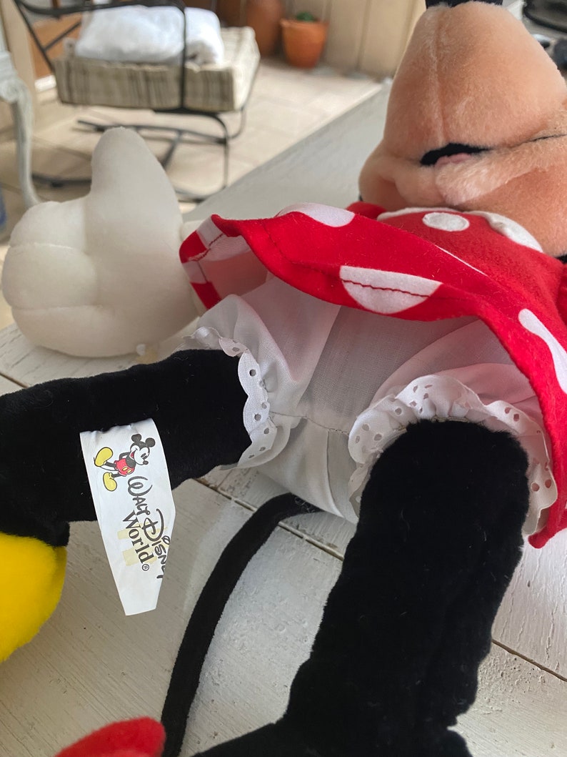 Może przedstawiać: Czerwono-biała sukienka w kropki z białym halką i czarnymi butami. Sukienka ma białą koronkową lam&oacute;wkę. Lalka nosi czarną opaskę na głowę z kokardką. Lalka ma ż&oacute;łtą twarz i czarne oczy. Lalka trzyma białą poduszkę w kształcie serca. Lalka ma metkę z napisem "Walt Disney World".