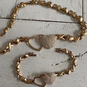 May include: A gold-tone necklace and bracelet set with interlocking heart charms. The hearts are adorned with sparkling rhinestones.