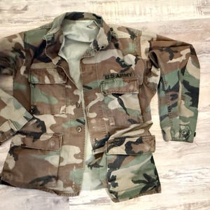 May include: Camouflage pattern jacket with multiple pockets and the text "U.S. ARMY" on the chest. The jacket features a button-up front and long sleeves. The color palette includes shades of brown, green, and tan.