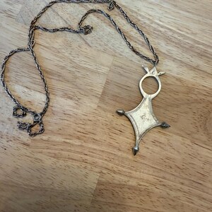 May include: A silver necklace with a pendant in the shape of a cross with a circle in the center. The pendant has a decorative design and pointed edges.