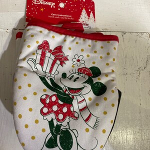 May include: A white oven mitt with a red border. The mitt features a cartoon image of Minnie Mouse holding a red and white gift box. Minnie Mouse is wearing a red dress with white polka dots, a white scarf with red stripes, and a red bow. The background is white with gold polka dots. The text "Care Instructions: Hand wash. Lay flat to dry." is printed on the packaging.