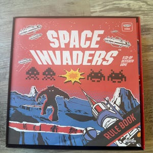 May include: A red and blue box with the words "Space Invaders" and "Rule Book" on it. The box features a retro video game design with a spaceship, aliens, and a monster.