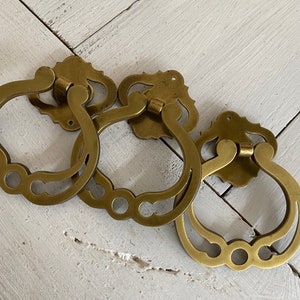 May include: Three ornate brass door pulls with a ring handle and decorative details. The pulls are arranged in a semi-circle on a white wooden surface.