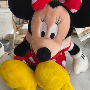 Może przedstawiać: Pluszowa zabawka Minnie Mouse w czerwonej sukience w białe kropki i czerwoną kokardkę w białe kropki. Zabawka ma ż&oacute;łte stopy i czarny nos. Zabawka ma metkę z napisem "Walt Disney World".