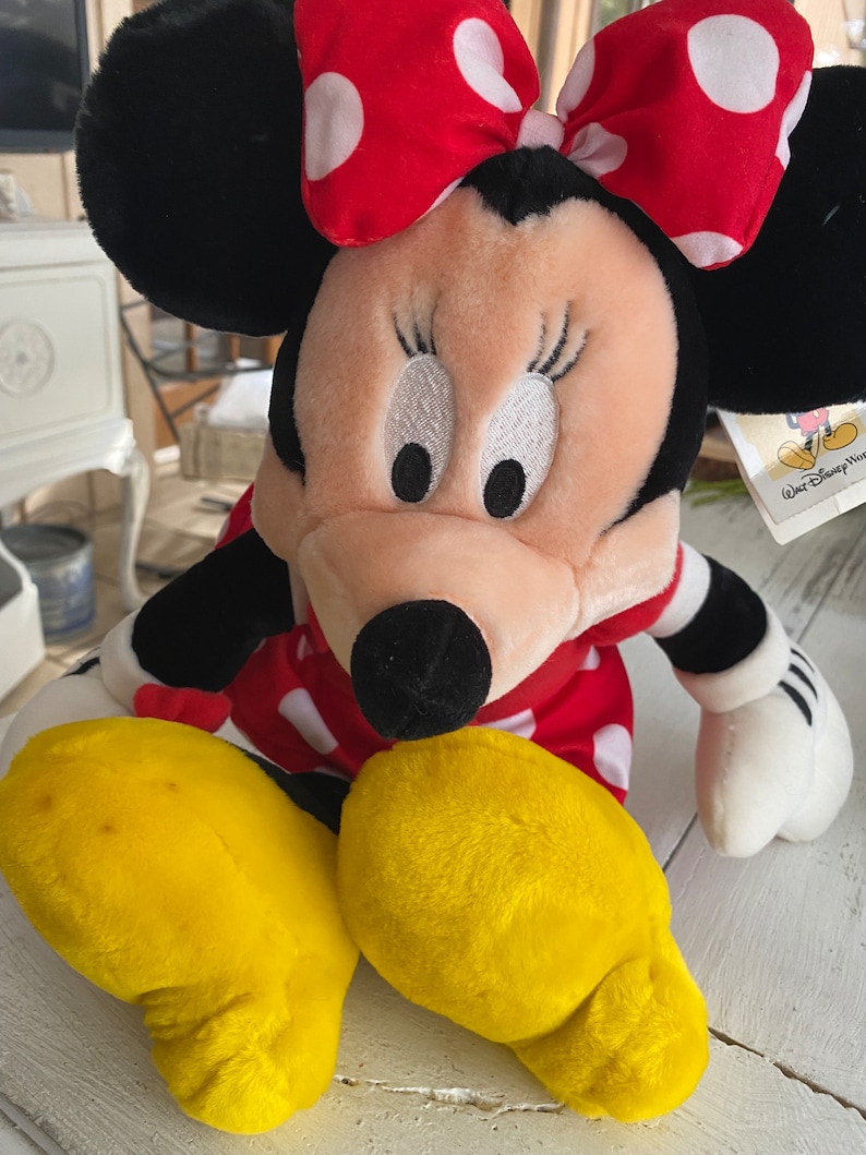 Może przedstawiać: Pluszowa zabawka Minnie Mouse w czerwonej sukience w białe kropki i czerwoną kokardkę w białe kropki. Zabawka ma ż&oacute;łte stopy i czarny nos. Zabawka ma metkę z napisem "Walt Disney World".