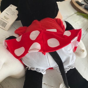 Może przedstawiać: Czerwona sukienka w białe kropki, z białą halką i czarnymi butami. Sukienkę nosi pluszowa zabawka Minnie Mouse z czarnymi uszami i czarnym ogonem.