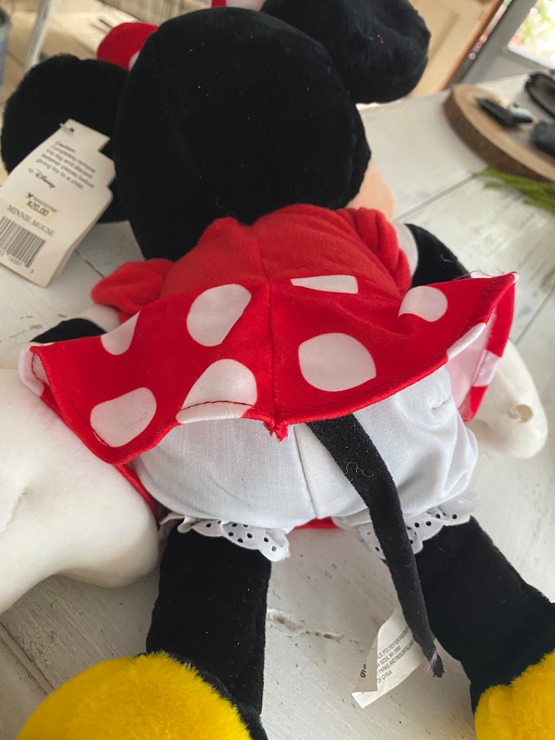 Może przedstawiać: Czerwona sukienka w białe kropki, z białą halką i czarnymi butami. Sukienkę nosi pluszowa zabawka Minnie Mouse z czarnymi uszami i czarnym ogonem.