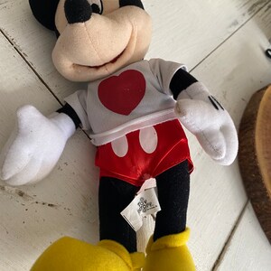 Może przedstawiać: Pluszowa zabawka Mickey Mouse ubrana w białą koszulkę z czerwonym sercem i czerwone spodenki w białe kropki. Zabawka ma czarne oczy, czarny nos i szeroki uśmiech. Zabawka siedzi na białym tle.