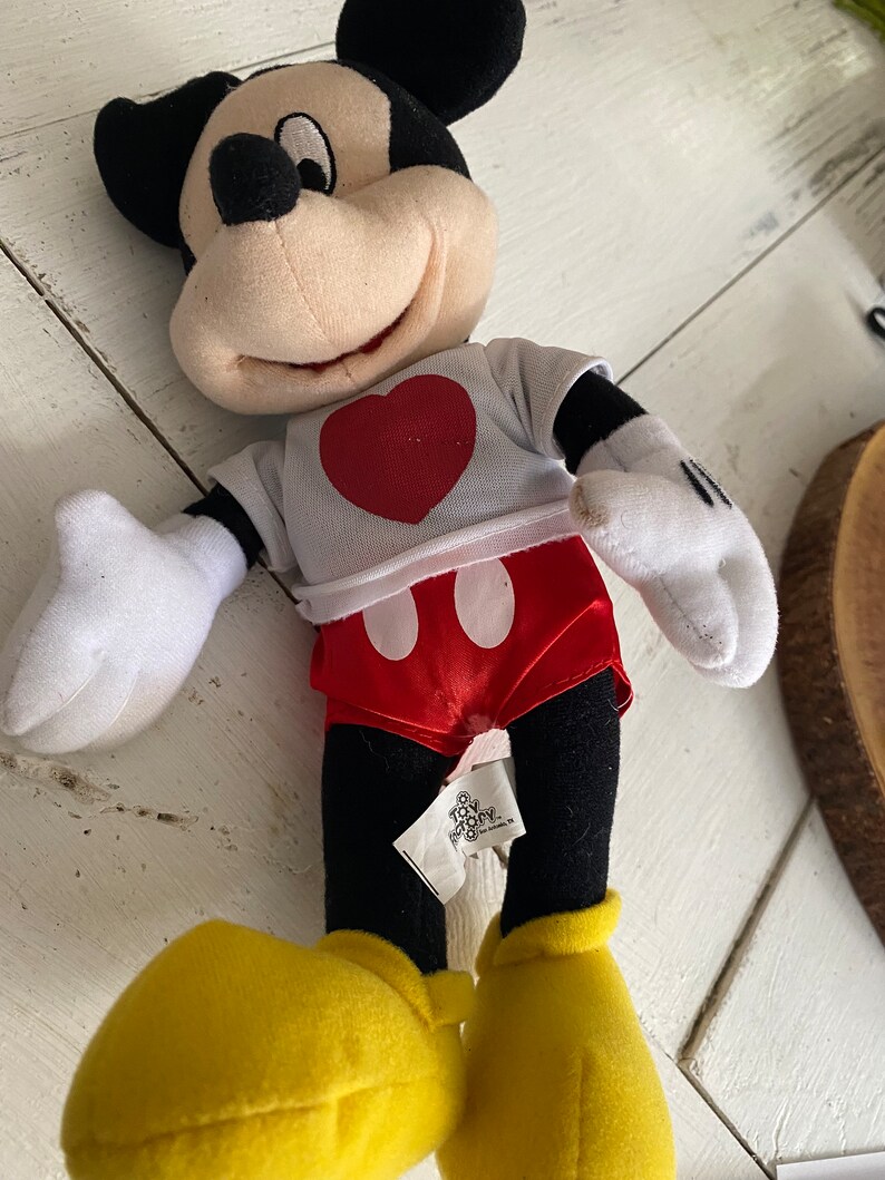 Może przedstawiać: Pluszowa zabawka Mickey Mouse ubrana w białą koszulkę z czerwonym sercem i czerwone spodenki w białe kropki. Zabawka ma czarne oczy, czarny nos i szeroki uśmiech. Zabawka siedzi na białym tle.