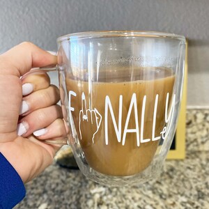 Könnte beinhalten: Eine doppelwandige Tasse aus klarem Glas mit einem weißen Design, auf dem "ENDLICH" mit einem Ringfinger und einem Ring steht. Die Tasse ist mit Kaffee gefüllt.