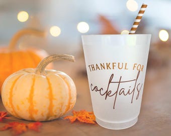 Op de afbeelding: Een matglazen kunststof beker met de tekst THANKFUL FOR cocktails in goudkleurige en bruine letters. Een bruin-wit gestreept papieren rietje zit in de beker. De beker staat naast een kleine, lichtgekleurde pompoen.