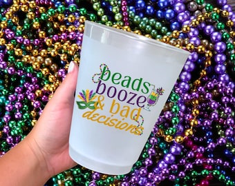 May include: A frosted plastic cup with the words "beads booze & bad decisions" in green, purple, and gold. The cup is held in a hand against a background of colorful Mardi Gras beads in green, gold, and purple.