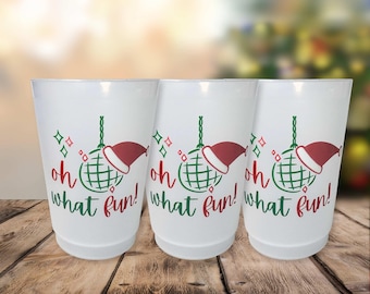 May include: Three white plastic cups with a festive design. Each cup features a green disco ball wearing a red Santa hat, with the words "oh what fun!" in green and red. The cups are on a wooden surface.