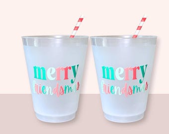 May include: Two frosted plastic cups with the words "merry friendsmas" in teal, pink, and red. Each cup has a red and white striped straw. The cups are on a light pink background.